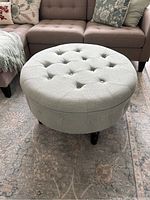 Front angled view of round light gray tufted fabric ottoman with button details on a patterned carpet, adjacent to sofa.