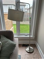 Full view of silver floor lamp standing near window and sofa, showing metal base, pole and white fabric lampshade.