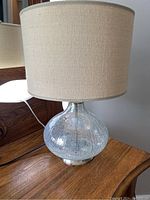 Full view of the glass table lamp on a wooden surface. Clear crackle glass base with beige fabric shade.