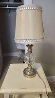 Full view of the lamp showing beige fabric shade with crystal trim and glass/metal base on a cream-colored table.