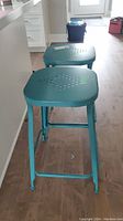 Two blue metal stools viewed from an angle, showing the square perforated seats and leg structure.