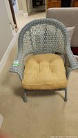 Front view of a grey wicker chair with high back and beige cushion placed on a carpeted floor.