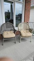 Two wicker chairs with beige tufted cushions on a porch.