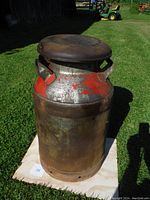 Antique milk can standing on grass showing worn red paint and rust patina from a front angle