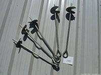 Six metal ground screw anchors for car shelters or tents, laid out on a light-colored surface, showing spiral tips and looped handles.