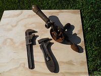 Photo showing two antique adjustable pipe wrenches and one portable hand drill on plywood board outdoors with grass background.