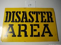 Photo of worn yellow 'DISASTER AREA' sign lying flat with visible surface wear and dirt marks.