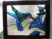 Close-up of the rectangular framed faux stained glass panel depicting a hummingbird and blue flower, wooden frame with chain hanging.