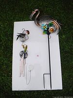Photo showing both items lying on a white board on grass: metal garden stake flamingo with colorful flowers and flamingo wind chime with glass globe and metal chimes.