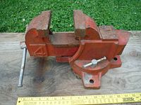 Full side view of the red metal bench vise on a wooden board, showing the large jaws, handle, and swivel base.
