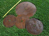 Four large, heavily rusted circular saw blades displayed on grass with a measuring stick showing size differences.