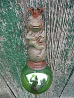 Close-up of cast iron frog figurine with crown and spiral eyes on green glass orb with reflective surface, textured weathered metal with rust patina.