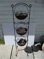 Full view of the black metal three-tier plant stand with scrollwork, showing the three baskets, soil residue, and sturdy frame.