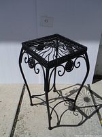 Full view of the black metal plant stand showing its square top with radiating bar design and decorative scrollwork on legs.