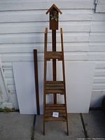 Full view of wooden plant stand shelf against white wall, showing three slatted shelves and decorative top with dried flower.