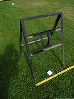 Metal sandwich sign frame standing on grass with measuring tape indicating size. Frame is black with two legs and crossbars, showing dirt and some wear.