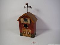 Front view showing barn-door painted front, circular entrance hole, hinged shutter with painted grass design, and weather vane rooster on roof.