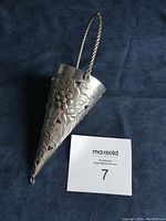 Full view of the conical metal bouquet holder with embossed floral design and twisted handle against dark background with a MaxSold lot card