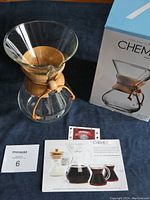 Chemex coffee maker with wooden collar, leather tie, original box and pamphlet on dark cloth.