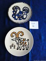 Two mid-century ceramic plates placed on a blue background, one with a stylized horse and one with a stylized goat, beside a lot card with number 18
