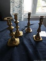 Four solid brass candleholders arranged on blue surface near window, showing two pairs with different designs and bases