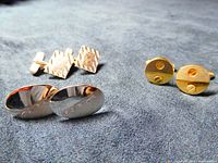 Three pairs of cufflinks shown on blue fabric: two pairs sterling silver, one pair golden and silver metallic Guy Laroche. Two pairs with distinct shapes and textures, third pair round with screw-like design.