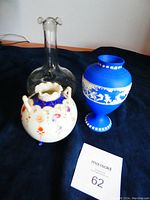 Three antique vases on a dark fabric background including white floral china vase, clear glass vase, and blue Jasperware vase