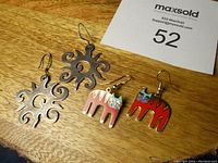 Four earrings arranged on wood surface with auction lot card Superimposed showing two enamel Laurel Burtch cat earrings in front and two sterling silver sunburst cutout earrings behind.