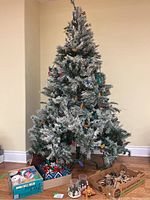 Full view of 7 ft snow covered artificial Christmas tree with lights, bows, and candle holders affixed on branches. Tree base and additional decorations visible underneath.