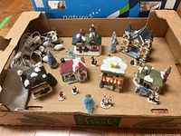 Overview image showing entire collection of 6 ceramic village houses, multiple figurines, trees, and string lights in a cardboard box.