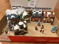 Wide view of all items including 5 ceramic houses, multiple small figurines, small Christmas trees, and light strand all arranged in a box.