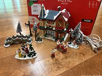 Full lot of ceramic Christmas village items arranged on floor, including toy shop building, carollers, trees, bridge, snowman, and lights with box in background