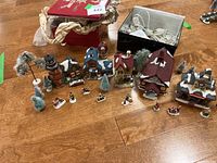 Top-down view showing the entire Christmas village set with ceramic houses arranged alongside trees, figurines, and a box containing string lights.