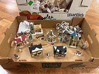 Overview of ceramic Christmas village lot in cardboard box showing multiple houses, trees, and figurines arranged together