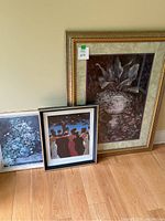 Three framed decorative art prints leaning against a wall: largest floral print in large gold frame, Renoir floral print, and Vettriano dance scene poster.