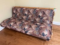 Front view of the futon sofa, showing the patterned brown floral fabric upholstery and wooden slat frame.