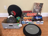 Photo shows box holding various games Dare, Master Mind, Masterpiece, Super Quiz, Crossword/Dominoes, Mindtrap plus sports balls and a Senators cushion.