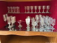Wooden cabinet with red velvet back containing about 50+ clear glass stemware pieces of various sizes and shapes including goblets, wine glasses, champagne flutes, and cocktail glasses.