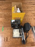 Kodak Instamatic M60 movie projector, original box, instruction manual, tripod, Mobilite Bounce Movie Light carrying case and cords laid out on wooden floor.