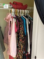 Clothing rack with pink puffer vest, floral and striped tops, and some dark colored skirts hanging on hangers in a closet.