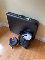 Folded black portable massage table with handle, detachable padded headrest, and striped carrying bag for sheets on hardwood floor