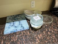 Photo of 3 ceramic casserole dishes with lids and extra lids stacked, plus one rectangular tempered glass cutting board with blue and white decorative design.