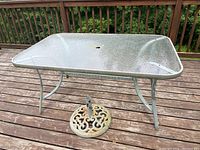 Photo of outdoor patio table with textured tempered glass top and metal frame showing rust and wear on legs and base, placed on wooden deck.