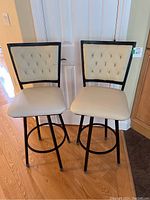 Pair of off-white pleather swivel bar stools with tufted backs, black metal frames, and circular footrests showing overall view.