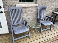 Two folding grey resin patio chairs with armrests positioned on wooden deck against stone wall beside door. Small round metal side table with glass top between chairs.