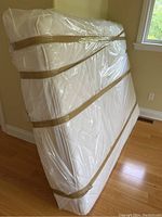 Side angle of queen mattress wrapped in clear plastic with securing straps, showing thickness and overall condition