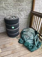 Stack of 4 tires with rims on porch next to green tire covers, showing overall set.