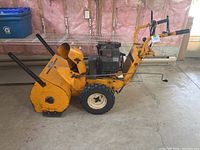 Side view of the yellow snow blower showing large rear tires, engine housing, handles, and chute for snow discharge.