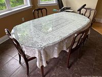 Dark wood veneer oval dining table with glass top protector and lace-patterned tablecloth set with 4 chairs around it, placed on tiled floor near windows.