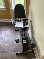 Stationary bike positioned against wall with seat, backrest, pedals, and handlebars visible. Owner's manual displayed on seat.
